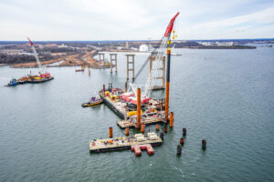 Barges support a crane in the water as it helps install construction piles. Old bridge supports seen in background.