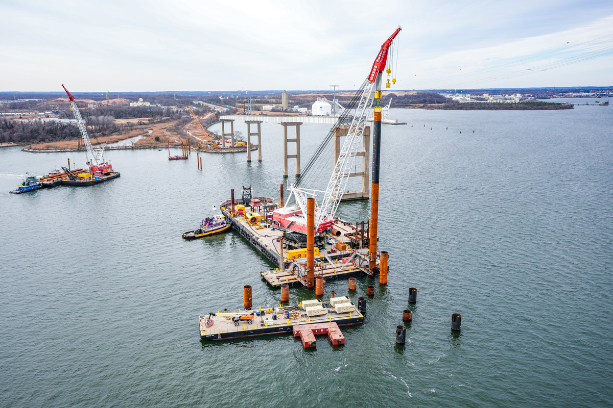 Barges support a crane in the water as it helps install construction piles. Old bridge supports seen in background.