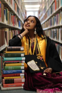 A college student wearing graduation regalia sits between two library shelfs leaning on a stack of books