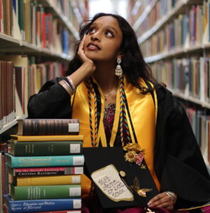 A college student wearing graduation regalia sits between two library shelfs leaning on a stack of books