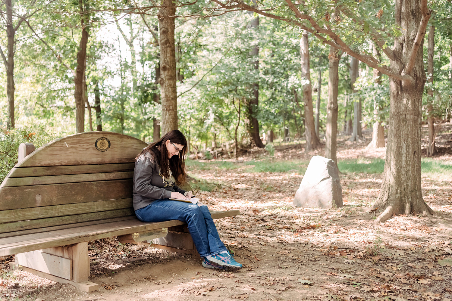 A person sits on a wooden bench in a wooded area, writing in a notebook.