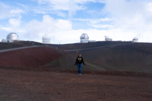 Morgan Robbins, wearing a UMBC sweatshirt, stands on barren rolling hills, five observatories visible in the background