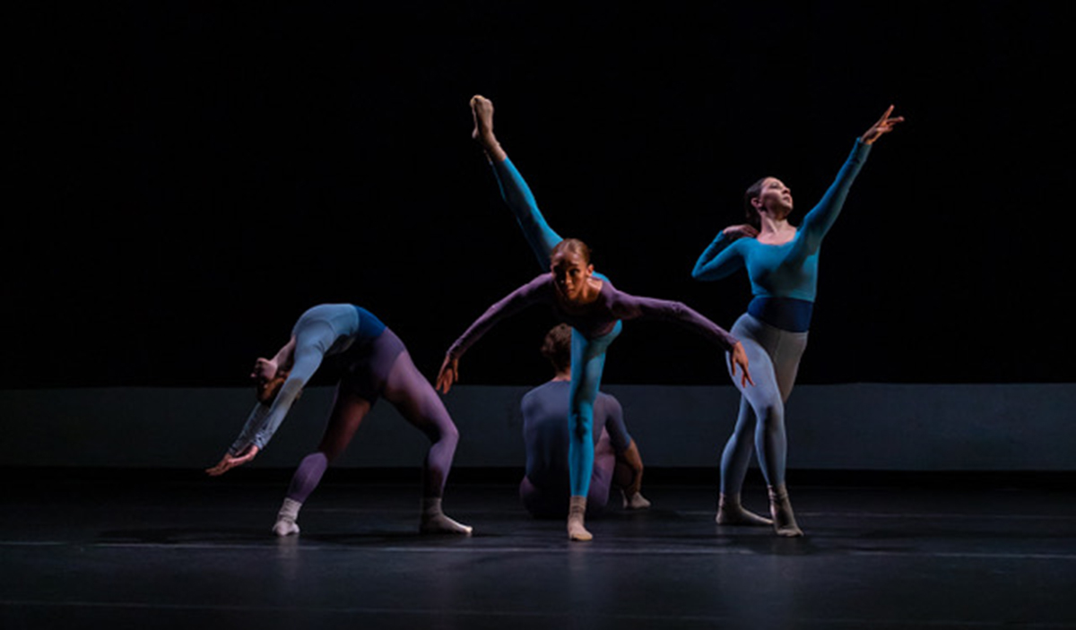 Four dancers perform a contemporary dance routine on a dark stage.