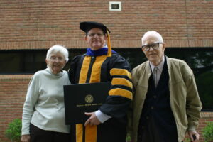 Man in center holds diploma and wears academic regalia. An older woman stands on his right and an older man on his left.