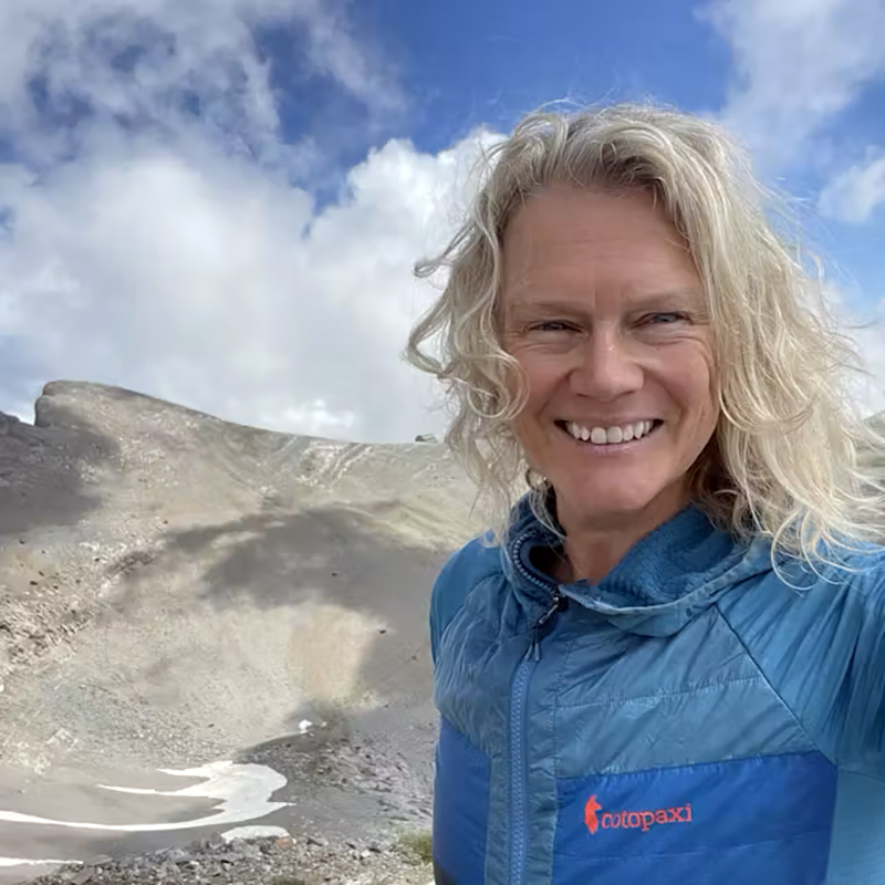 A person smiles while standing on a mountain slope with rocky terrain and patches of snow.