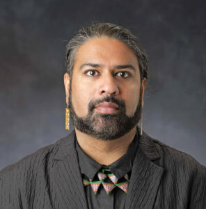 Headshot of a person with salt and pepper hair, a beard, and a nose piercing, wearing a textured black jacket and a collar with geometric patterns.
