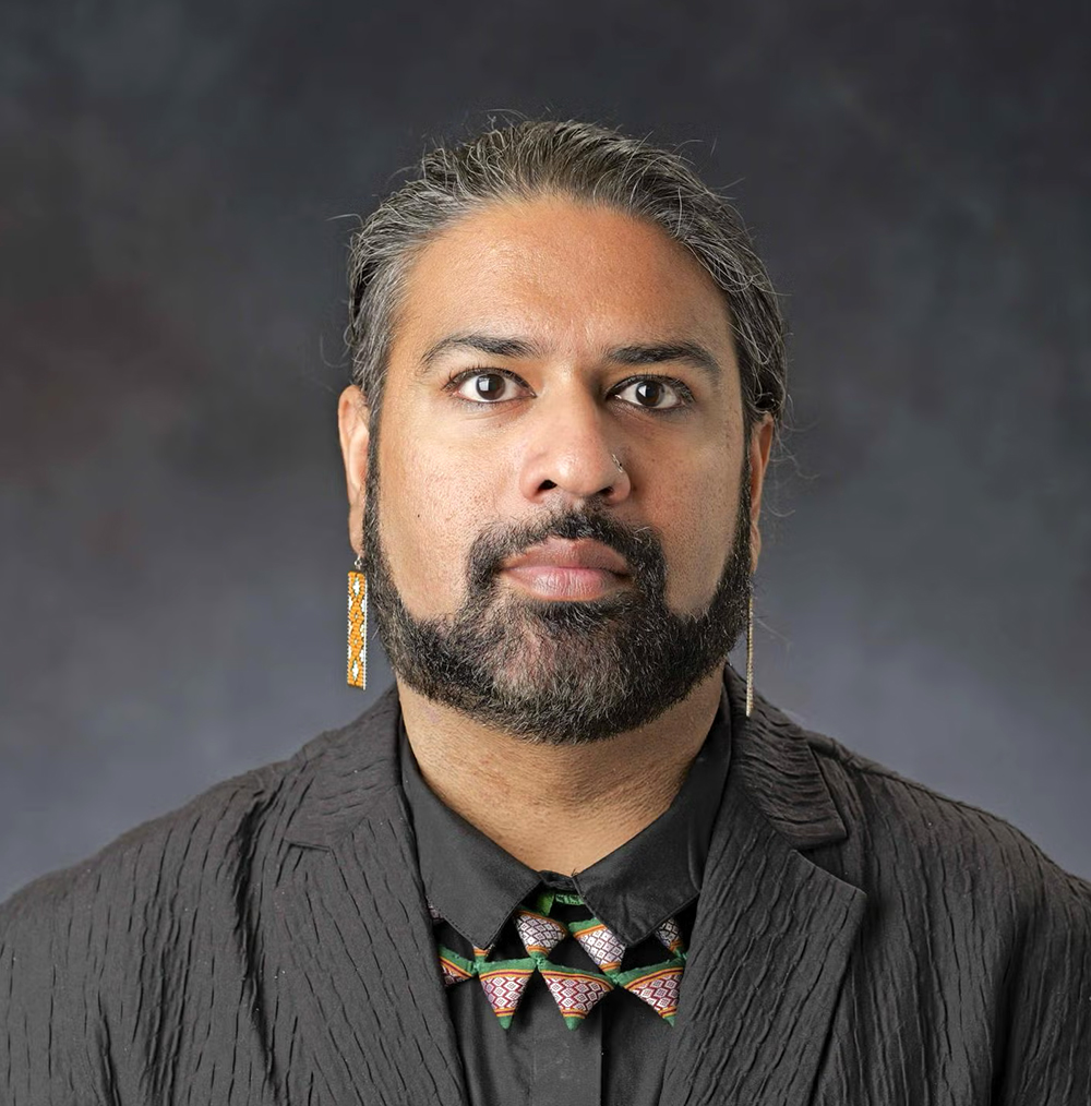 Headshot of a person with salt and pepper hair, a beard, and a nose piercing, wearing a textured black jacket and a collar with geometric patterns.
