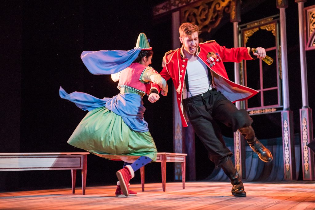 Two performers in theatrical costumes dance on stage, with one in a bright uniform jacket and the other in a full skirt and headpiece.