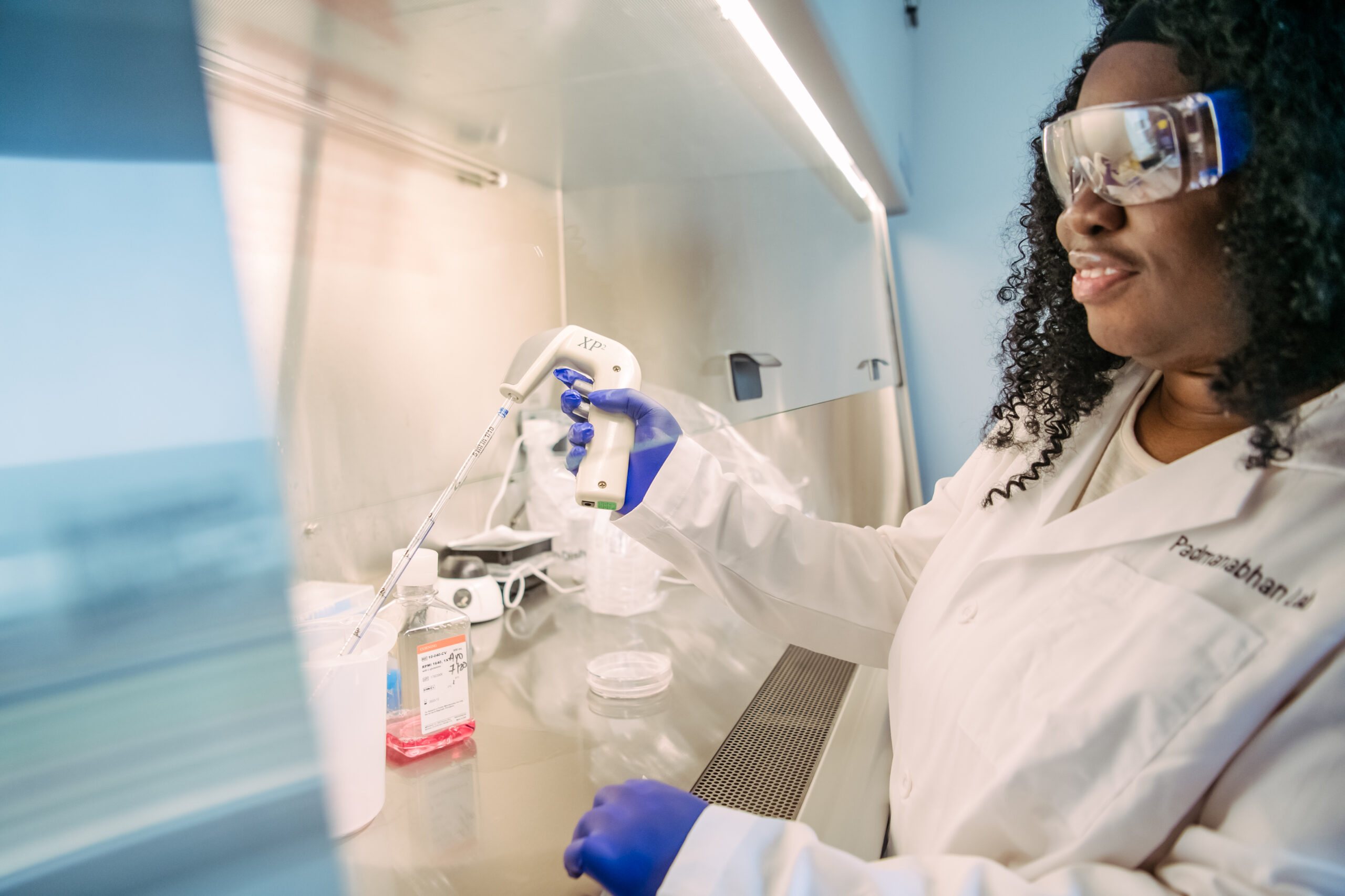 Ayo Ogunsanya works at a fume hood, in lab coat, goggles, and gloves, pipetting into a flask with pink liquid