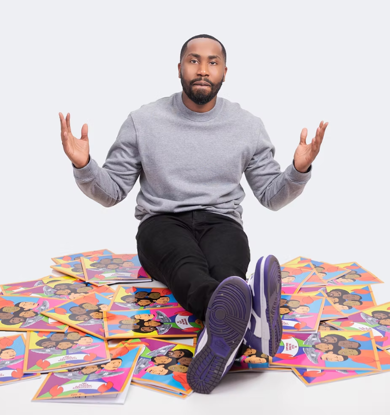 Devin Wilkins wearing black jeans and a grey sweatshirt sitting on top of a pile of the Black Baltimore Renaissance Coloring Books