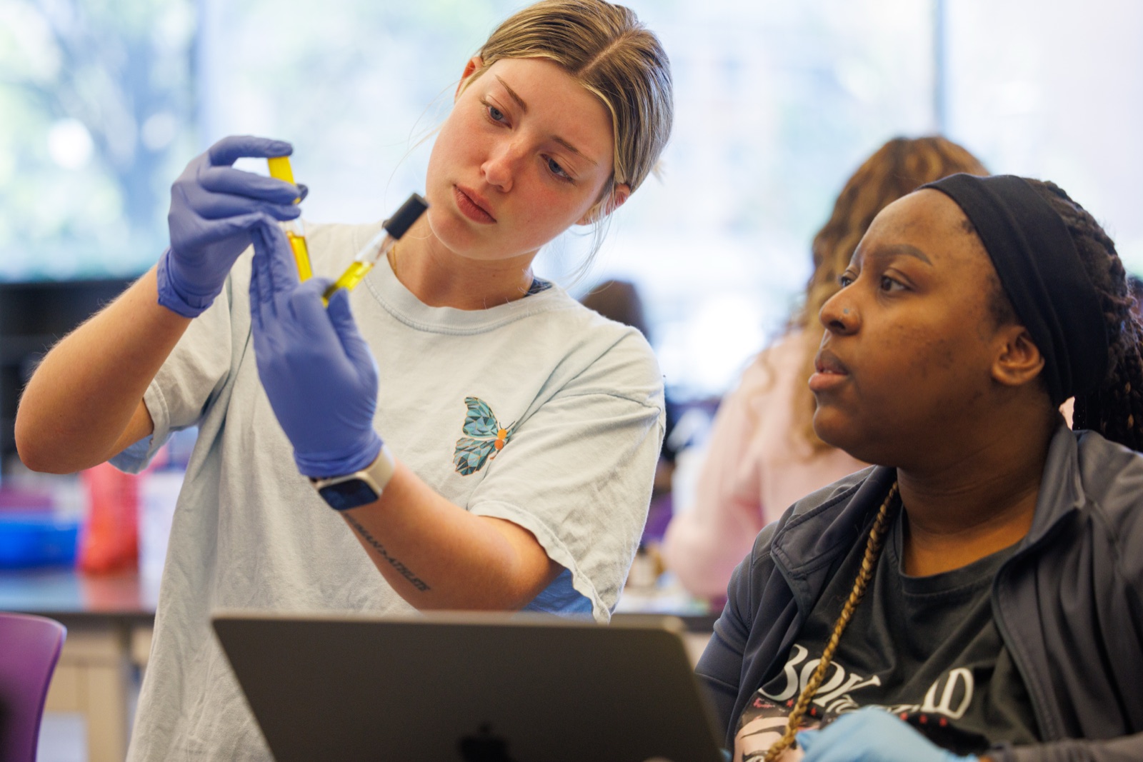 Two students wearing gloves examine two yellow liquid samples in a laboratory setting while working with a laptop.