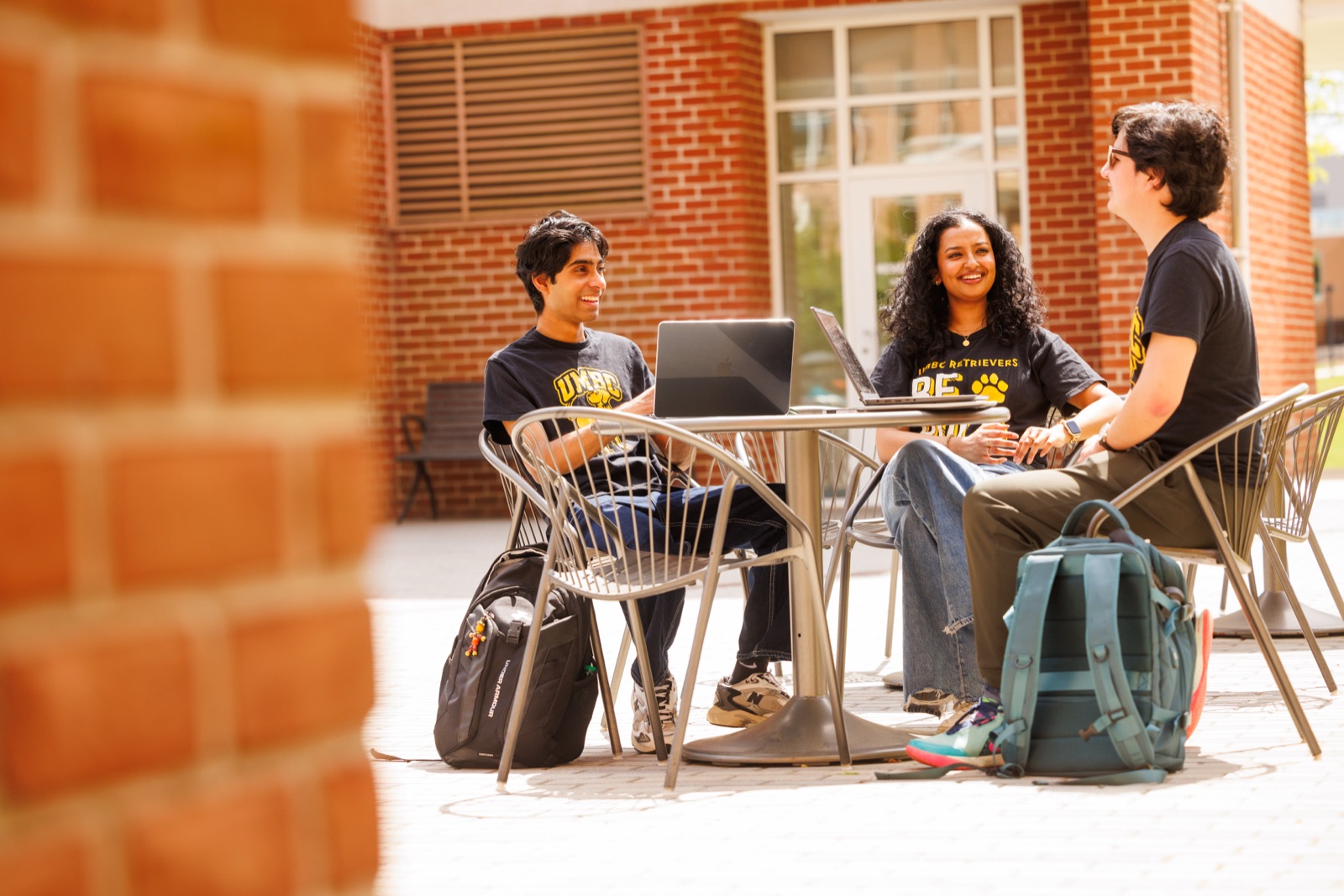 Three students sit at an outdoor table with laptops, talking and smiling.