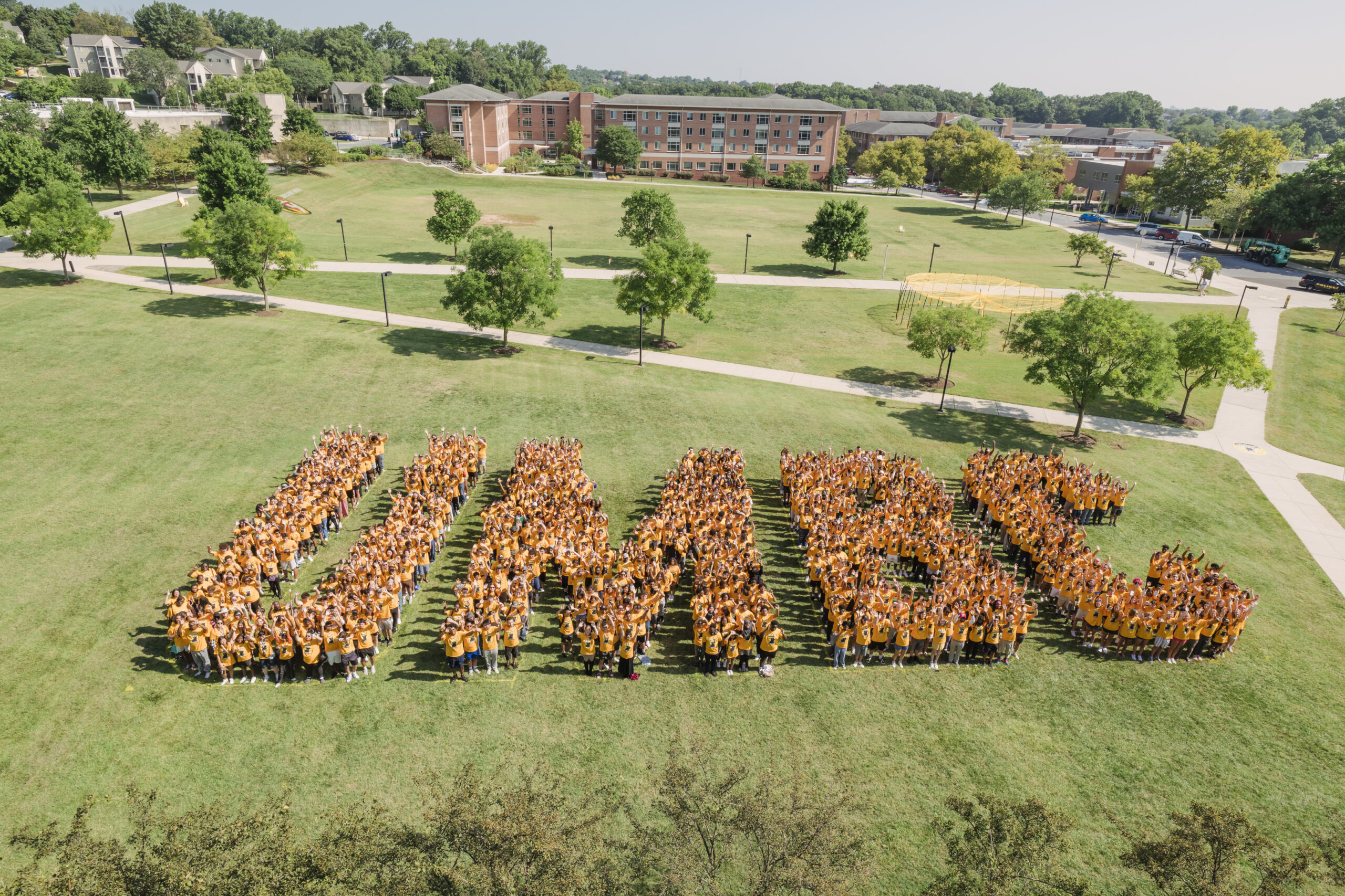 A large group of students in matching yellow shirts stand on a grassy field, arranged to form the letters UMBC while waving their arms.